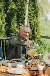 Senior man pouring juice in glass sitting at dining table - VSNF00812