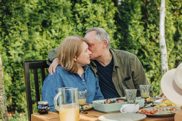 Älterer Mann küsst Frau, die am Esstisch im Hinterhof sitzt - VSNF00813