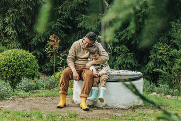 Vater mit Sohn sitzen zusammen auf einem Brunnen im Garten - VSNF00815