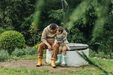Sohn flüstert Vater zu, der im Garten am Brunnen sitzt - VSNF00817