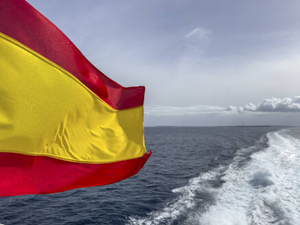 Spain, Balearic Islands, Formentera, Wake left by moving boat with Spanish flag fluttering in foreground - MMAF01489