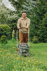 Smiling man standing with lawnmower in garden - VSNF00820