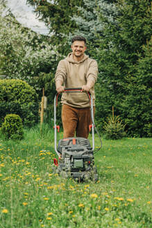 Smiling man standing with lawnmower in garden - VSNF00820