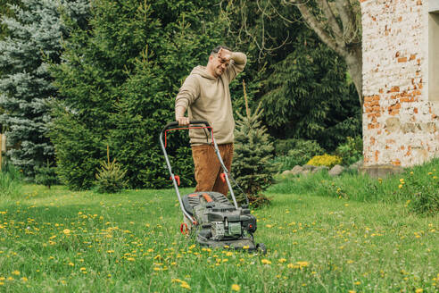 Tired man standing by lawnmower in garden - VSNF00821