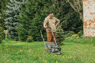 Mann macht Pause mit Rasenmäher im Garten stehend - VSNF00822