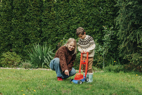 Mother teaching son to mow grass using toy lawnmower in garden - VSNF00824