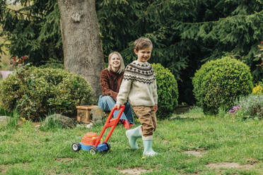 Mutter beobachtet ihren Sohn beim Spielen mit einem Spielzeugrasenmäher im Garten - VSNF00825