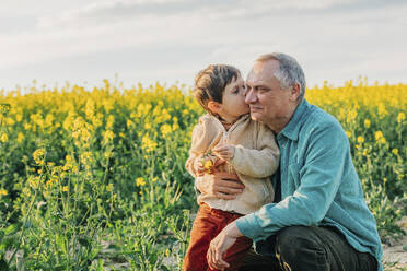 Grandson kissing grandfather crouching in rapeseed field - VSNF00838