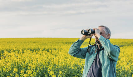 Senior man looking through binoculars amidst rapeseed field - VSNF00843