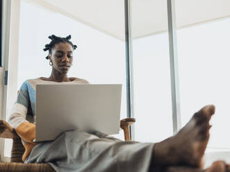 Young woman using laptop sitting comfortably in armchair at home - MFF09333