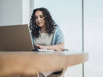 Eine entschlossene Frau mit lockigem Haar arbeitet konzentriert an ihrem Laptop am Schreibtisch - MFF09334