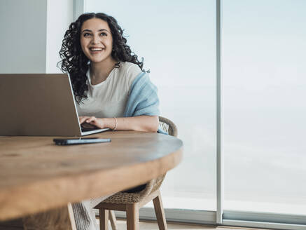 Eine fröhliche Frau, die gemütlich zu Hause sitzt, vertieft in ihre Arbeit am Laptop, lächelt etwas aus dem Off an - MFF09335
