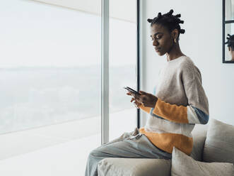 Young woman using mobile phone sitting on sofa at home - MFF09341