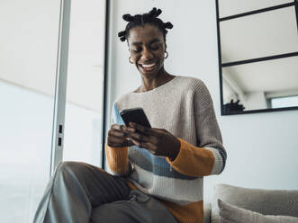 Smiling woman reading text messages on smartphone at home - MFF09342