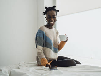 Eine fröhliche Frau, die ihren Morgenkaffee genießt, während sie auf ihrem Handy surft und sich im Bett entspannt - MFF09353