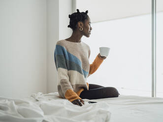 Woman sitting on bed holding coffee cup looking out of window - MFF09354