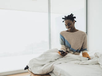 Cheerful woman sitting on bed looking at screen of smartphone - MFF09357