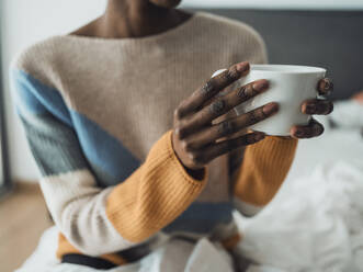 Eine Frau sitzt auf einem Bett und genießt ihren Morgenkaffee, während sie die Tasse mit beiden Händen hält - MFF09363