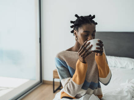 Eine junge Frau genießt eine Tasse Kaffee, während sie gemütlich in ihrem Schlafzimmer sitzt - MFF09364