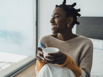 Smiling woman sitting on bed holding coffee cup - MFF09367