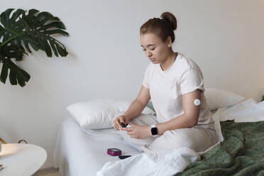 Young woman with diabetes checking glucose level sitting on bed at home - AAZF00463