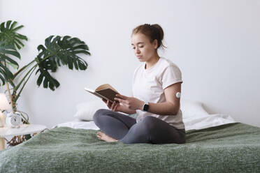 Frau mit Diabetes liest ein Buch im Schneidersitz auf dem Bett zu Hause - AAZF00528