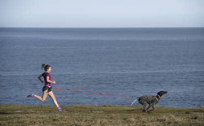 Woman running with dog on grass near sea - SNF01636