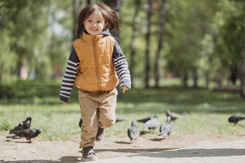 Fröhlicher Junge läuft inmitten von Tauben im Park - ANAF01410