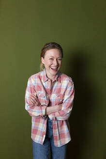 Junge Frau in kariertem Hemd lacht mit verschränkten Armen vor einer grünen Wand - MIKF00338