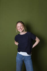 Lächelnde junge Frau in blauem T-Shirt steht mit den Händen hinter dem Rücken an einer grünen Wand - MIKF00341