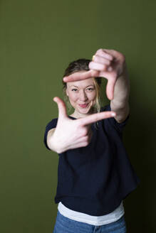 Smiling young woman in blue T-shirt gesturing finger frame in front of green wall - MIKF00342