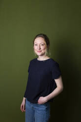 Confident young woman in blue T-shirt standing with hands in pockets in front of green wall - MIKF00343