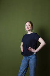 Confident young woman in blue T-shirt standing with arms akimbo in front of green wall - MIKF00345