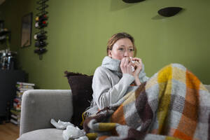 Kranke junge Frau putzt sich die Nase mit einem Taschentuch und entspannt sich auf der Couch - MIKF00348
