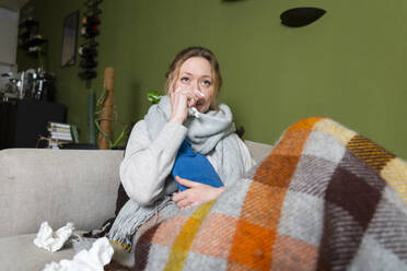 Kranke junge Frau wischt sich auf der Couch die Nase mit einem Taschentuch ab - MIKF00350