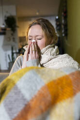 Sick young woman wrapped in blanket sneezing at home - MIKF00352