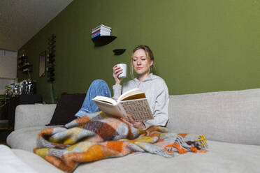 Junge Frau mit Tasse und Buch auf der Couch - MIKF00358