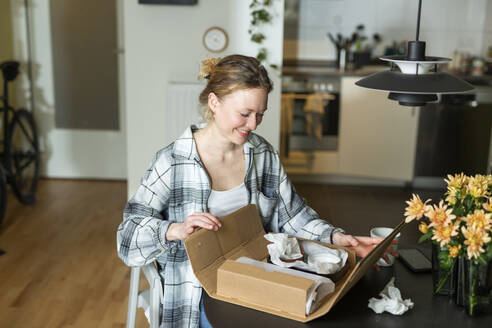 Smiling young woman unpacking ceramic bowls from cardboard box on table - MIKF00375