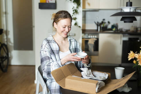 Smiling young woman unpacking ceramic bowls from cardboard box at home - MIKF00376
