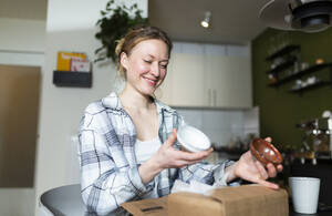 Smiling young woman unpacking ceramic bowls from cardboard box - MIKF00377