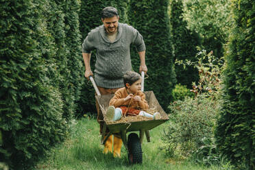 Father pushing son sitting on hay in wheelbarrow at back yard - VSNF00888