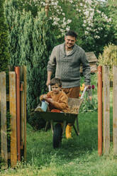 Father pushing son sitting in wheelbarrow at back yard - VSNF00891