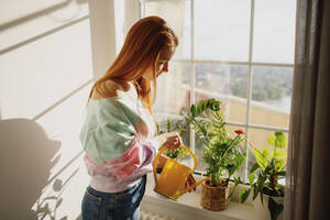 Rothaarige Frau bewässert blühende Pflanzen auf der Fensterbank zu Hause - MDOF01105