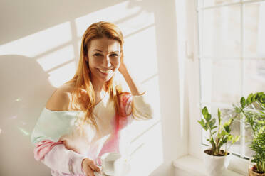 Smiling redhead woman with coffee cup standing by wall at home - MDOF01111