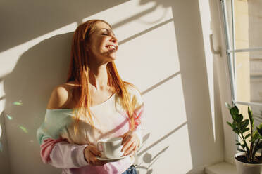 Lächelnde rothaarige Frau mit Kaffeetasse genießt das Sonnenlicht am Fenster zu Hause - MDOF01112