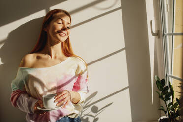 Redhead woman with coffee cup enjoying sunlight by window at home - MDOF01113