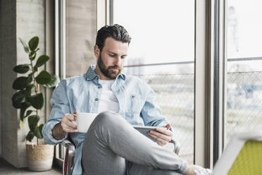 Businessman using tablet PC sitting with coffee cup at office - UUF28588