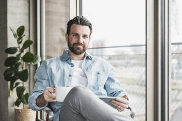 Smiling businessman sitting with coffee cup at office - UUF28589