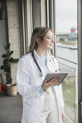Female doctor looking out of window holding tablet PC at hospital - UUF28599