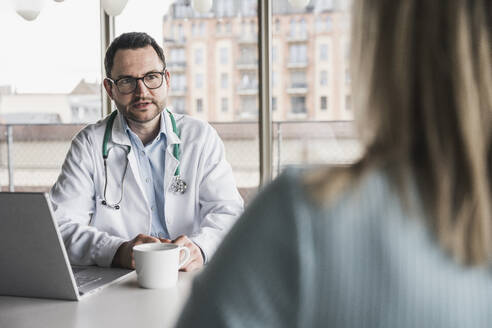 Arzt berät Patientin am Schreibtisch sitzend in der Klinik - UUF28602